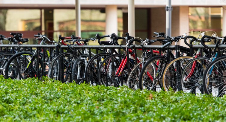 Full view of a bycicle parking, filled with bikes, in front of a green garden.