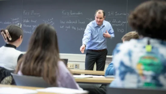 A male professor teaching a class in front of his students facing him and the blackboard.
