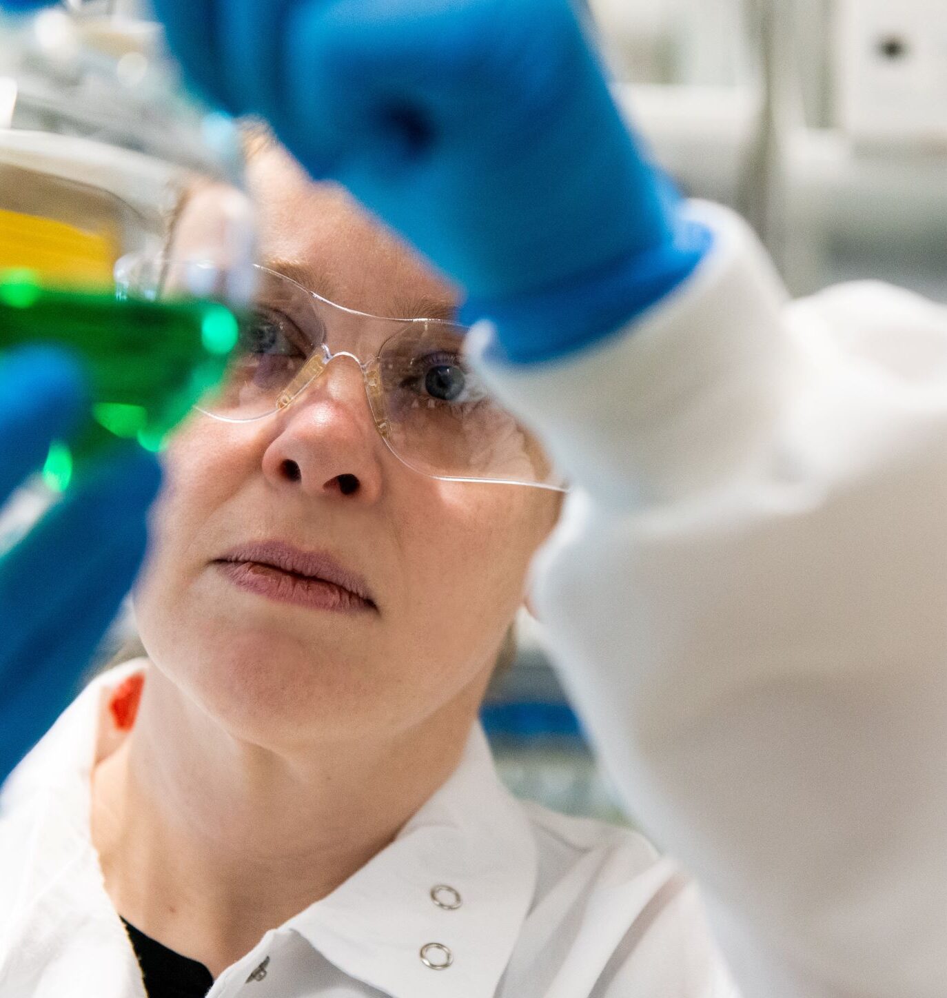 Woman wearing a white lab coat and blue latex gloves inspecting a beaker containing a green liquid solution