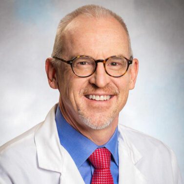 Portrait of Gregg Greenough. Portrait of a man with white hair, wearing glasses and a white lab coat.