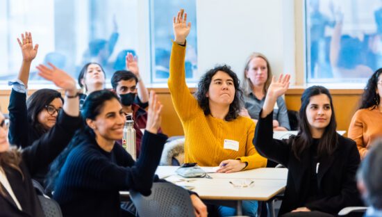 Six students sitting in the audience at an event raise their hands.