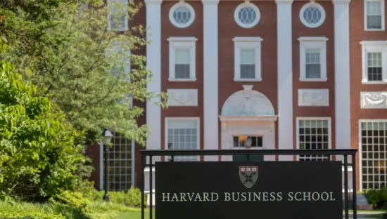 A sign reads Harvard Business School. Behind the sign, a brick building with white columns stands.