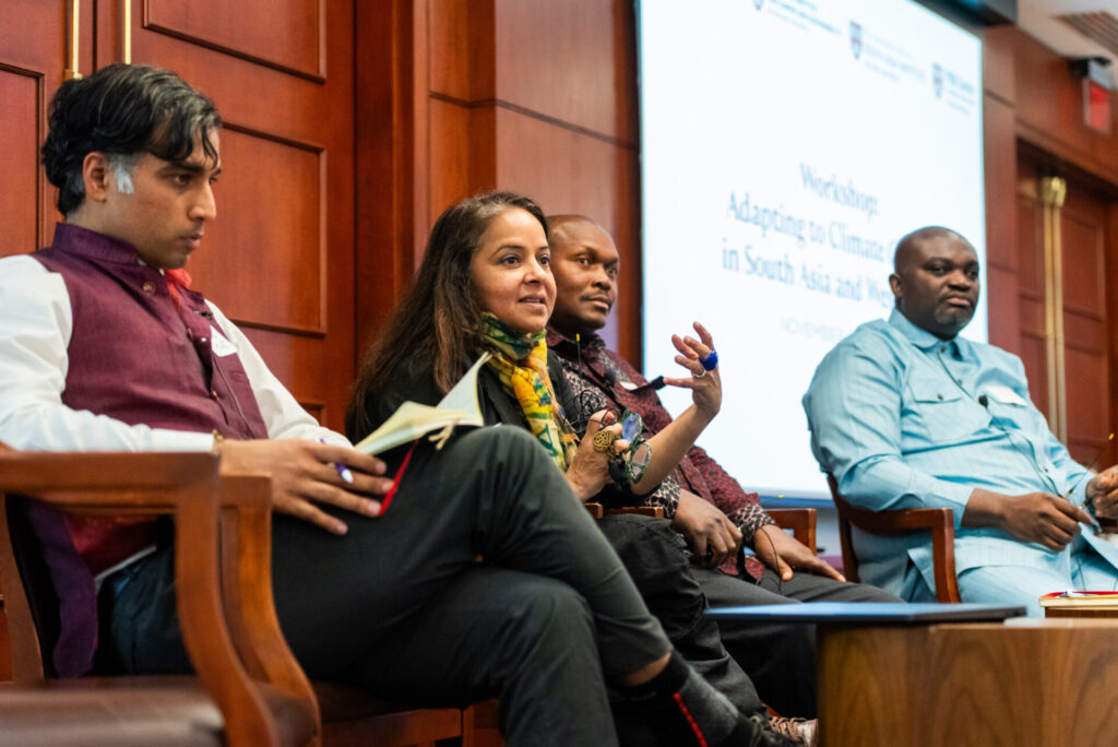 A person gestures while speaking, surrounded by three other panelists. A screen behind the speakers reads "workshop: Adapting to Climate Change in South Asia and West Africa."