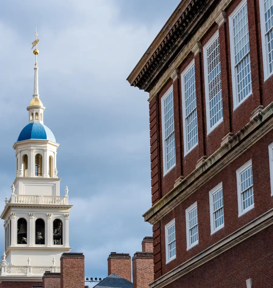 Picture of Harvard buildings, on the left there is the top of a white spire with a blue room that is Lowell house, on the right a brick building with tall windows.