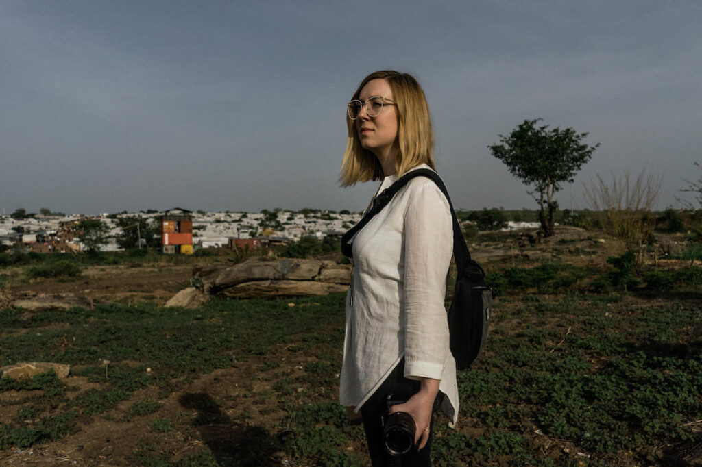 Journalist Denise Hruby stands in a a field with low-lying structures in the background. She holds a camera.