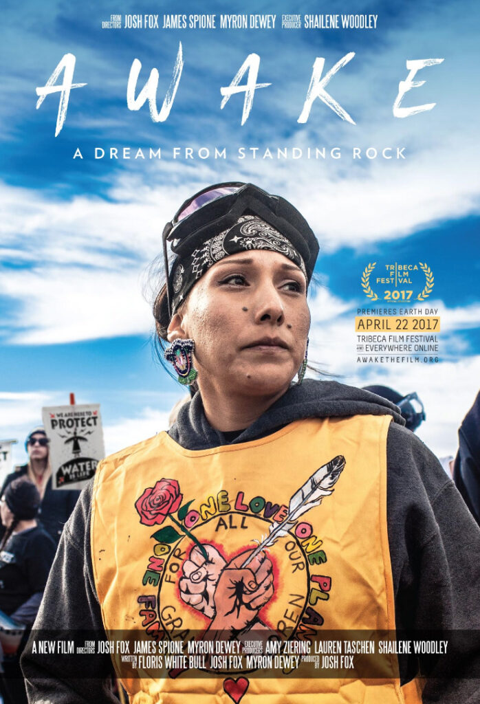 A person wears a bandana, goggles on their head, with a yellow vest over a gray hoodie. The yellow best shows fists holding a rose and a feather. Awake: A Dream from Standing Rock 