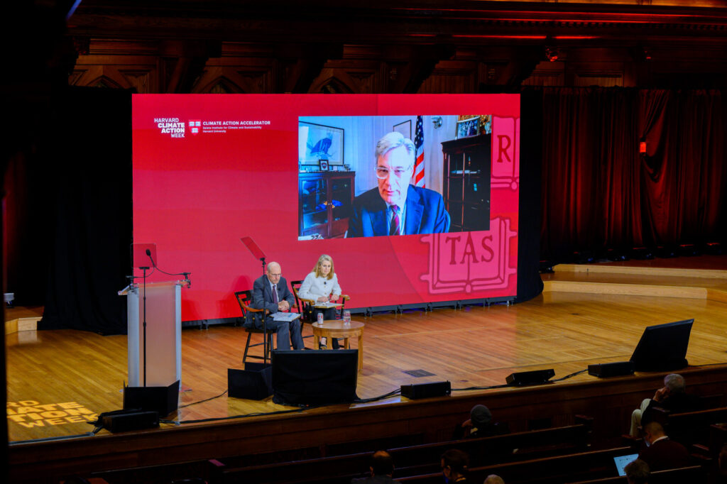 Senator Whitehouse appears on a crimson screen behind economists James Stock and Catherine Wolfram, seated on stage.